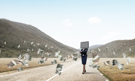 Business woman in suit with TV instead of head keeping arms crossed while standing on the road among flying dollar banknotes with beautiful landscape on background. の写真素材