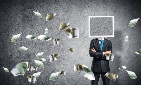 Businessman in suit with monitor instead of head keeping arms crossed while standing among flying euros and with gray dark wall on background.の写真素材