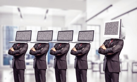 Businessmen in suits with laptops instead of their heads keeping arms crossed while standing in a row and one at the head with monitor inside office building.の写真素材