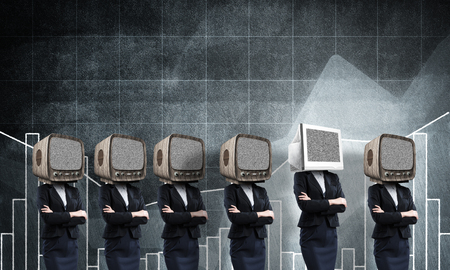 Business women in suits with old TV instead of their heads keeping arms crossed while standing in a row and one at the head with monitor against analytical charts on background.の写真素材