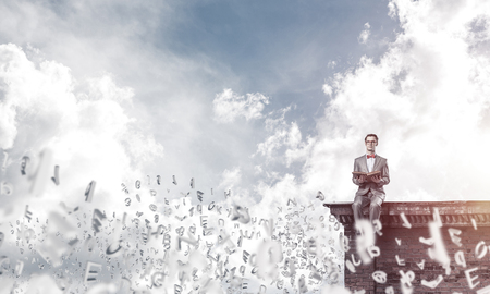 Funny man in red glasses and suit sitting on building top and reading bookの写真素材