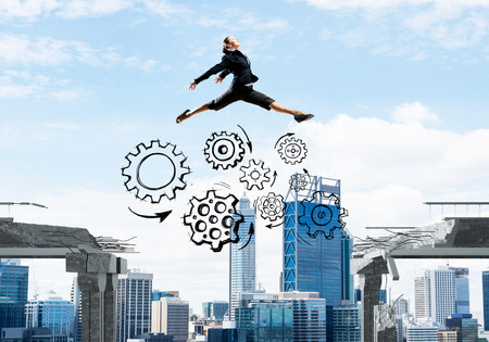 Business woman jumping over gap with gear mechanism in concrete bridge as symbol of overcoming challenges. Cityscape on background. 3D rendering.の写真素材
