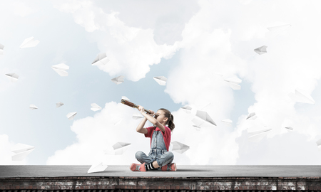 Cute kid girl sitting on house roof and looking in spyglassの写真素材