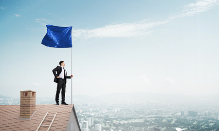 Businessman standing on house roof and holding blue flag. Mixed mediaの写真素材