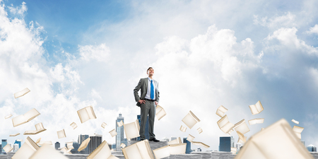 Confident businessman in suit standing on pile of documents among flying books with cloudly sky on background. Mixed media.の写真素材