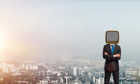 Cropped image of businessman in suit with old TV instead of head keeping arms crossed while standing outdoors with cityscape view on background.の写真素材