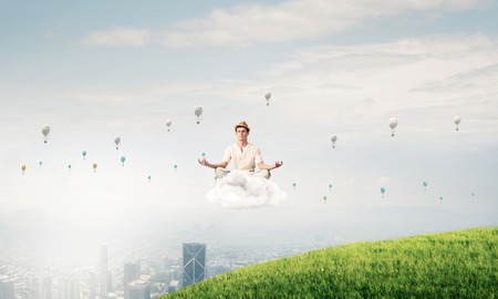 Young man in white clothing keeping eyes closed and looking concentrated while meditating on cloud in the air with city view on background.の写真素材