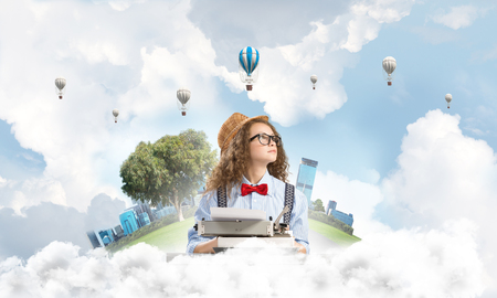 Young and beautiful woman writer in hat and eyeglasses using typing machine while sitting at the table with floating city island and cloudy skyscape with flying aerostats on background.の写真素材