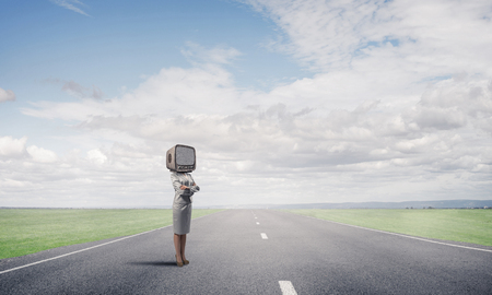 Business woman in suit with an old TV instead of head keeping arms crossed while standing on the road with beautiful landscape on background.の写真素材