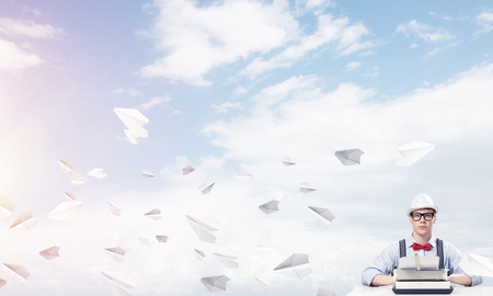 Young man writer in hat and eyeglasses using typing machine while sitting at the table among flying paper planes with cloudy skyscape on background.の写真素材