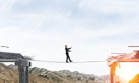 Businessman walking blindfolded on rope above huge gap in bridge as symbol of hidden threats and risks. Skyscape and nature view on background. 3D rendering.の写真素材