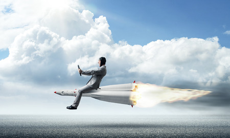 Conceptual image of young businessman in suit flying on rocket above asphalt road with blue cloudy sky on background.の写真素材
