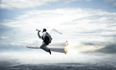 Conceptual image of young businessman in suit flying on rocket above asphalt road with blue cloudy sky on background.の写真素材