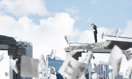 Young engineer in suit and helmet looking down while standing among flying papers on broken bridge with cityscape on background. 3D rendering.の写真素材