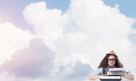 Young and beautiful woman writer in hat and eyeglasses using typing machine while sitting at the table with cloudy skyscape on background.の写真素材