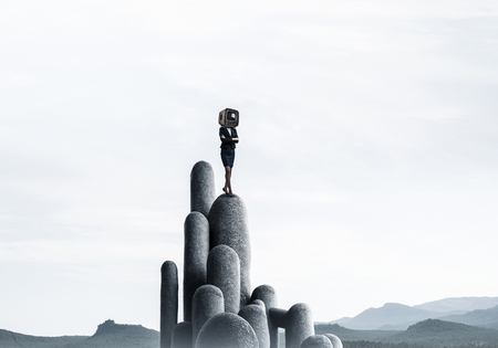 Business woman in suit with an old TV instead of head keeping arms crossed while standing on the top of stone columns with beautiful landscape on background.の写真素材