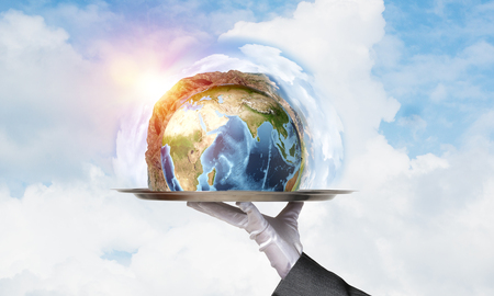 Cropped image of waitress's hand in white glove presenting Earth globe on metal tray with cloudy skyscape on background. 3D rendering. Elements of this image are furnished by NASA.の写真素材