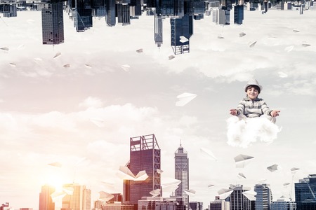 Young boy keeping eyes closed and looking concentrated while meditating on cloud among flying paper planes between two urban worlds.の写真素材