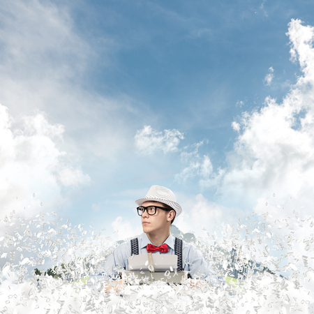 Young man writer in hat and eyeglasses using typing machine while sitting at the table among flying letters with floating city and cloudy skyscape on background.の写真素材