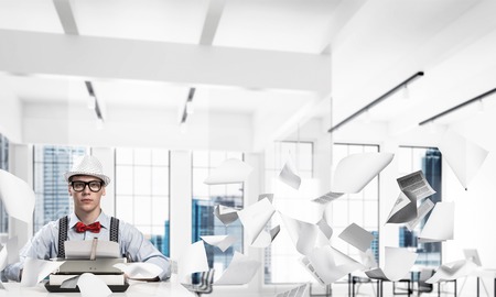 Young man writer in hat and eyeglasses using typing machine while sitting at the table indoors among flying papers and with office view on background.の写真素材
