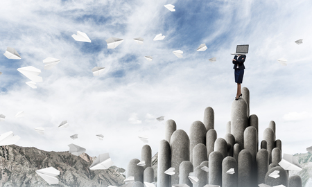 Business woman in suit with laptop instead of head keeping arms crossed while standing on the top of stone column with beautiful skyscape on background. 3D rendering.の写真素材