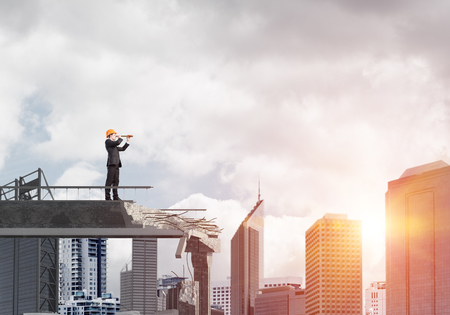 Young engineer in suit and helmet looking in spyglass while standing on broken bridge with cityscape and sunlight on background. 3D rendering.の写真素材