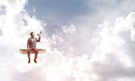 Young businessman sitting on book and screaming emotionally in megaphoneの写真素材