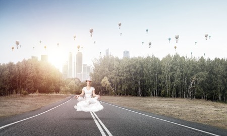 Young woman keeping eyes closed and looking concentrated while meditating on cloud above the road with beautiful and breathtaking landscape on background.の写真素材