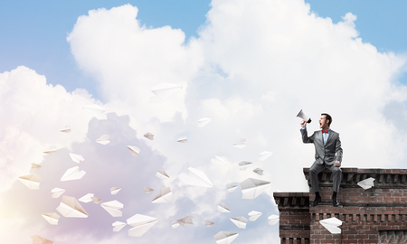 Young businessman sitting on roof and screaming emotionally in megaphoneの写真素材