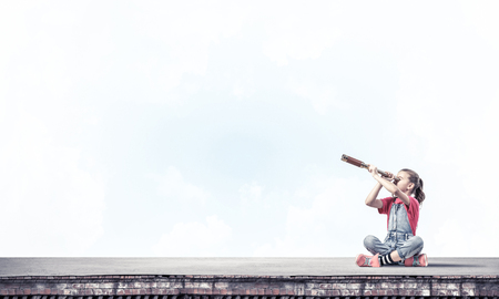 Cute kid girl sitting on house roof and looking in spyglassの写真素材