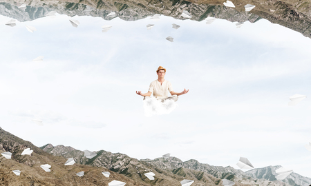 Young man keeping eyes closed and looking concentrated while meditating on cloud among flying paper planes and between two nature worlds.の写真素材