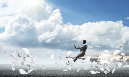Conceptual image of young businessman in suit flying on rocket above asphalt road and among flying papers with blue sky on background.の写真素材