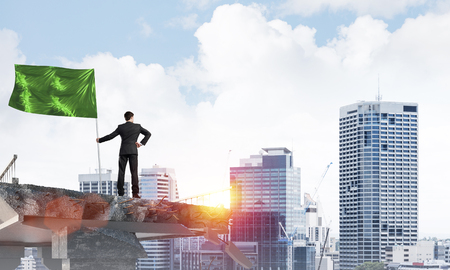 Rear view of confident businessman in suit holding green flag in hand while standing on broken bridge with cityscape and sunlight on background. 3D rendering.の写真素材