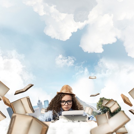 Young and beautiful woman writer in hat and eyeglasses using typing machine while sitting at the table among flying books with floating city island and cloudy skyscape on background.の写真素材