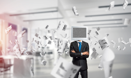 Cropped image of businessman in suit with monitor instead of head keeping arms crossed while standing among flying papers inside office building.の写真素材