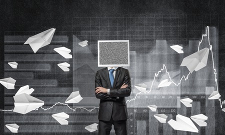 Businessman in suit with monitor instead of head keeping arms crossed while standing against flying paper planes and analytical charts drawn on wall on background.の写真素材