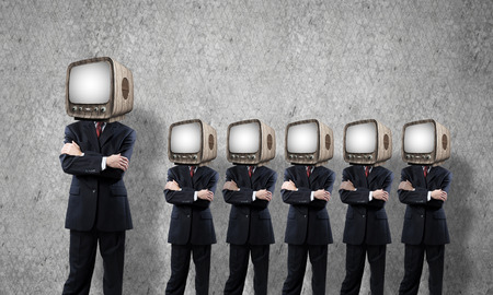 Businessmen in suits with old TV instead of their heads keeping arms crossed while standing in a row in empty room with gray wall on background.の写真素材