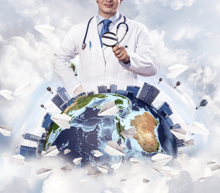 Cropped image of professional man doctor in white suit and stethoscope on neck discovering Earth globe through magnifier, with cloudy skyscape on background. Medical industry concept. Elements of this image furnished by NASAの写真素材