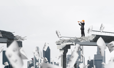 Young engineer in suit and helmet looking in spyglass while standing among flying papers on broken bridge with cityscape on background. 3D rendering.の写真素材