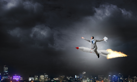 Conceptual image of young businessman in suit flying on rocket with night cityscape and blue sky on background.の写真素材