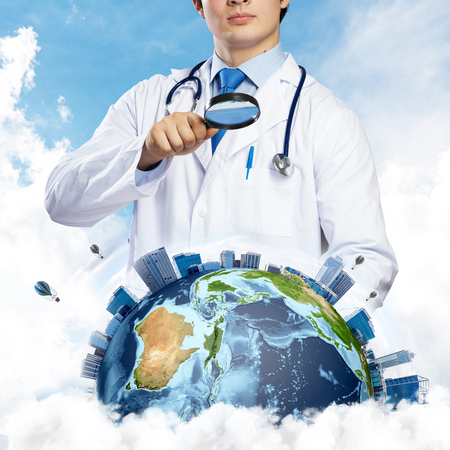 Cropped image of professional man doctor in white suit and stethoscope on neck discovering Earth globe through magnifier, with cloudy skyscape on background. Medical industry concept.の写真素材