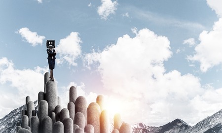 Business woman in suit with an old TV instead of head keeping arms crossed while standing on the top of stone columns with beautiful landscape on background.の写真素材