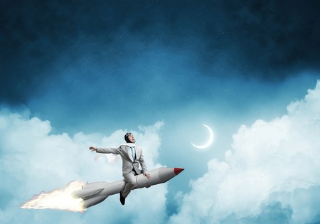 Conceptual image of young businessman in suit flying on rocket with blue night skyscape with clouds and moon on background.の写真素材