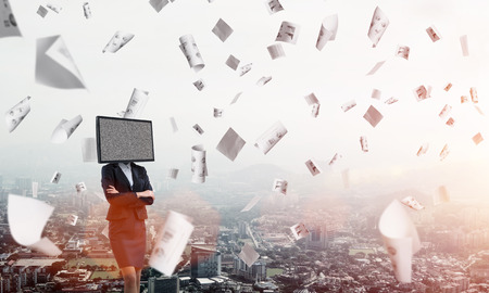 Cropped image of business woman in suit with TV instead of head keeping arms crossed while standing outdoors among flying papers with cityscape view on background.の写真素材
