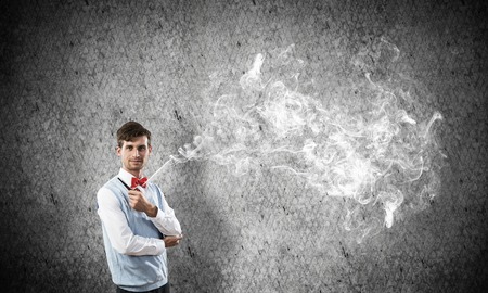 Horizontal shot of confident and young businessman in smart-casual wear smoking pipe while standing against gray dark wall on background.の写真素材