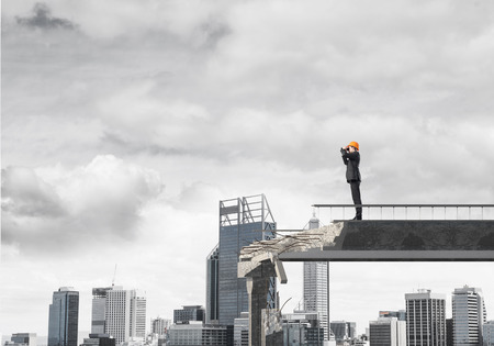 Young engineer in suit and helmet looking in binoculars while standing on broken bridge with cityscape on background. 3D rendering.の写真素材