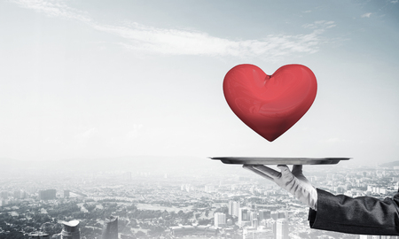 Cropped image of waitress's hand in white glove presenting big red heart on metal tray with cityscape view on background.の写真素材