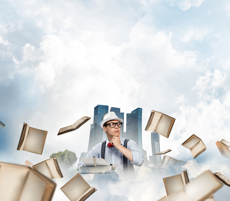 Young man writer in hat and eyeglasses using typing machine while sitting at the table among flying books with floating city and cloudy skyscape on background.の写真素材