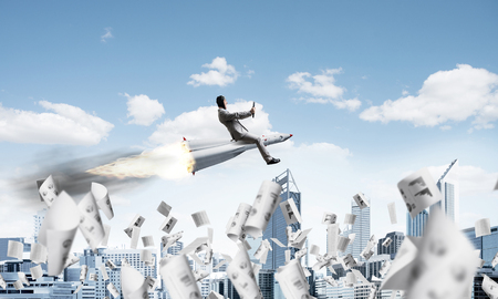 Conceptual image of young and happy businessman in suit flying on rocket among flying papers with modern cityscape with skyscrapers and blue sky on background.の写真素材