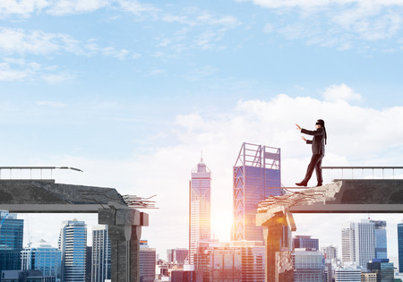 Businessman walking blindfolded on concrete bridge with huge gap as symbol of hidden threats and risks. Cityscape view with sunlight on background. 3D rendering.の写真素材